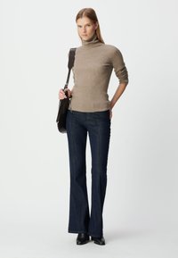 Taupe geribd coltrui en donkerblauwe uitlopende spijkerbroek. Model houdt een bruin leren tas vast. De laarzen zijn zwart met een spitse neus.