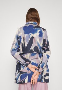 Henkilö, jolla on pitkä ruskea tukka ja p�äällään sinivalko-pinkki abstraktikuosiset pitkähihainen takki, kädet ristissä selän takana, katsoo poispäin.