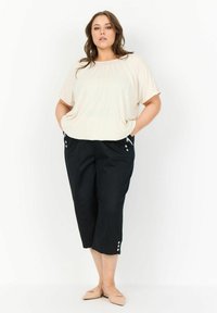 Béžový top s krátkym rukávom v kombinácii s čiernymi 3/4 nohavicami so bočnými vreckami a gombíkovými detailami; model stojí s rukami vo vreckách.