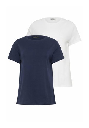 Kvinde med blondt hår iført en navyblå kortærmet skjorte, lyseblå vidde jeans og hvide sneakers, stående mod en ensfarvet baggrund.