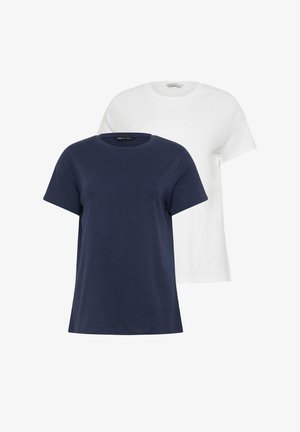 Vrouw met blond haar, een marineblauw T-shirt met korte mouwen, lichtblauwe wijde jeans en witte sneakers, staand tegen een effen achtergrond.