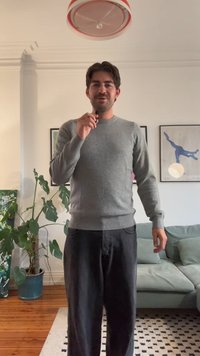 Hallukas halli kudum, pikkade varrukatega ja ümmarguse kaelusega, koos mustade vabalt istuvate pükstega, seistes möbleeritud eluruumis.