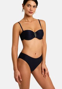 Femme portant un bikini deux pièces noir avec des bretelles fines et une fermeture décorative au centre du haut, debout devant un fond uni.