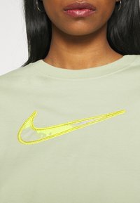 Světle zelené bavlněné tričko s výrazným vyšitým žlutým logem Nike s texturovaným designem. Detailní pohled na výstřih a logo.