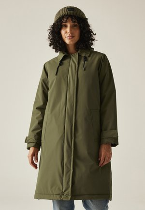 Kobieta z kręconymi włosami, ubrana w oliwkowozielony długi pikowany płaszcz i dopasowaną dzianinową czapkę, stojąca na tle jednolitego jasnego tła.