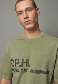 Oliven grøn bomulds t-shirt med rund hals, der har sort tekst og grafiktryk på fronten. Blød tekstur og afslappet pasform.
