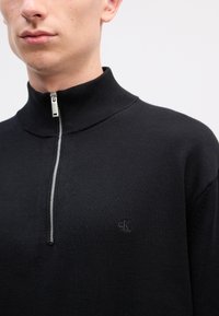 Czarny dzianinowy sweter zapinany na zamek błyskawiczny z wysokim kołnierzem, z metalowym zamkiem oraz małym tłoczonym logo na piersiach. Gładka, teksturowana tkanina.