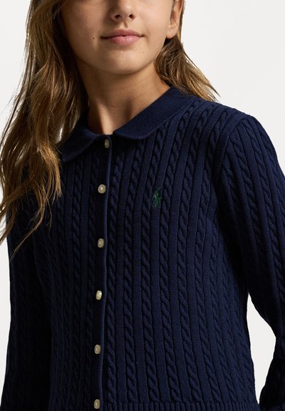 Ναυτικό μπλε πλεκτό πουλόβερ με σχέδιο καλωδίων, με γιακά και κουμπιά που �μοιάζουν με κοσμήματα, διακοσμημένο με ένα μικρό πράσινο λογότυπο στο στήθος.