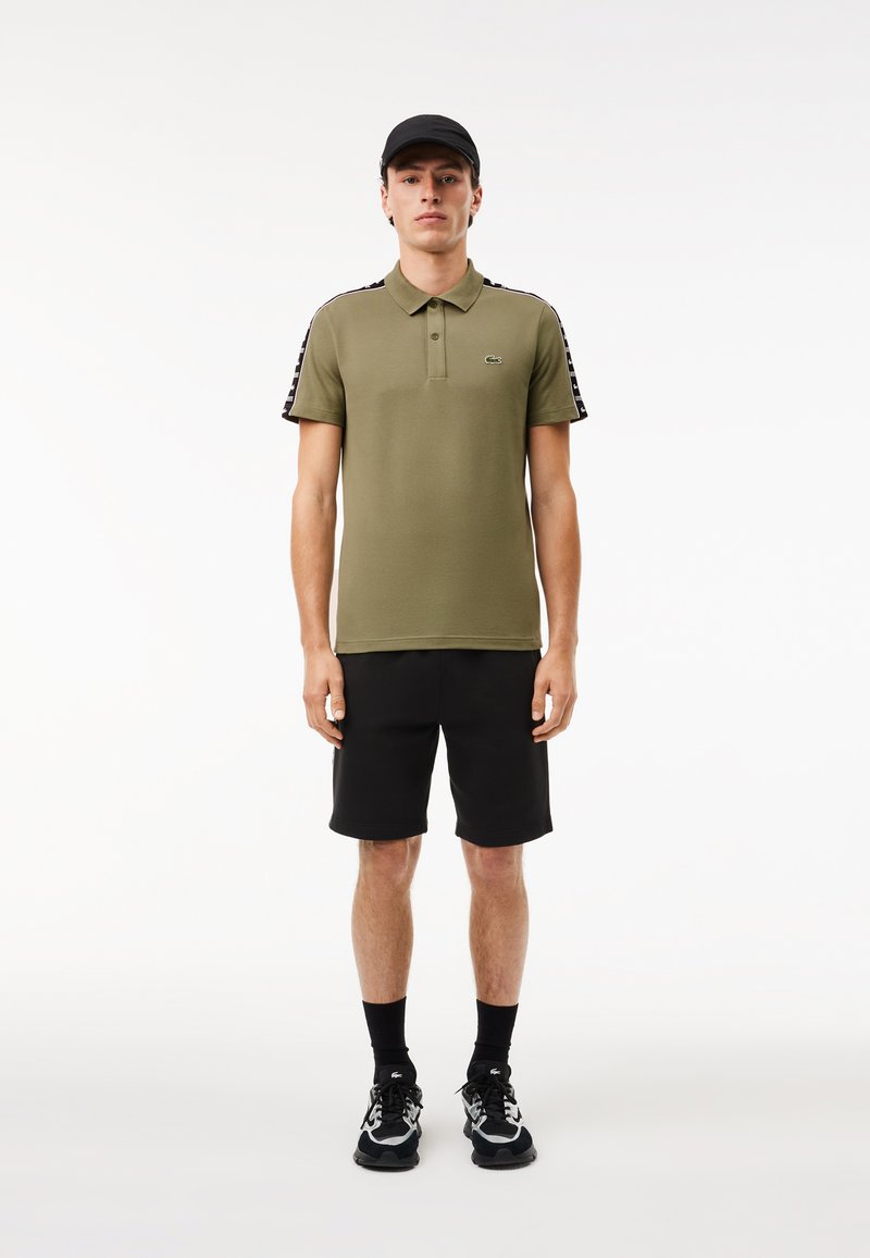 Polo vert olive avec épaules à rayures noires, shorts noirs et baskets noires. Texture lisse, col classique et détail de logo.