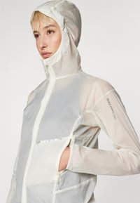 Lightweight, transparent hooded jacket in off-white, featuring a front zipper, reflective accents, and a side pocket. Textured fabric visible.