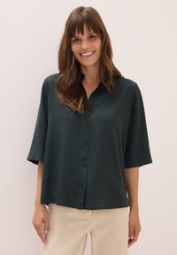 Kvinna som ler, iklädd en mörkgrön lös skjorta med knappar och ärmar som går till armbågarna, samt ljusbeige byxor, står framför en ljus bakgrund.