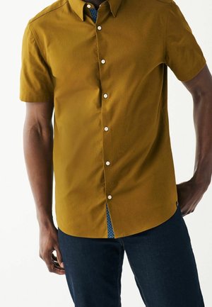 Man in een mosterdgele korteblouse met knopen over een donkerblauwe spijkerbroek, staand met een hand in de zak tegen een witte achtergrond.