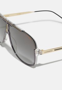 Ochelari de soare cu ramă transparentă și lentile cu gradient întunecat. Prezintă accente metalice aurii și branding pe bradă. Design unghiular cu un aspect modern.