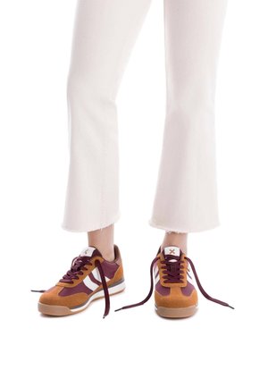 Zapatillas bajas de ante marrón y burdeos, con rayas blancas, cordones redondos y suela de goma. Complementan unos jeans blancos de campana.