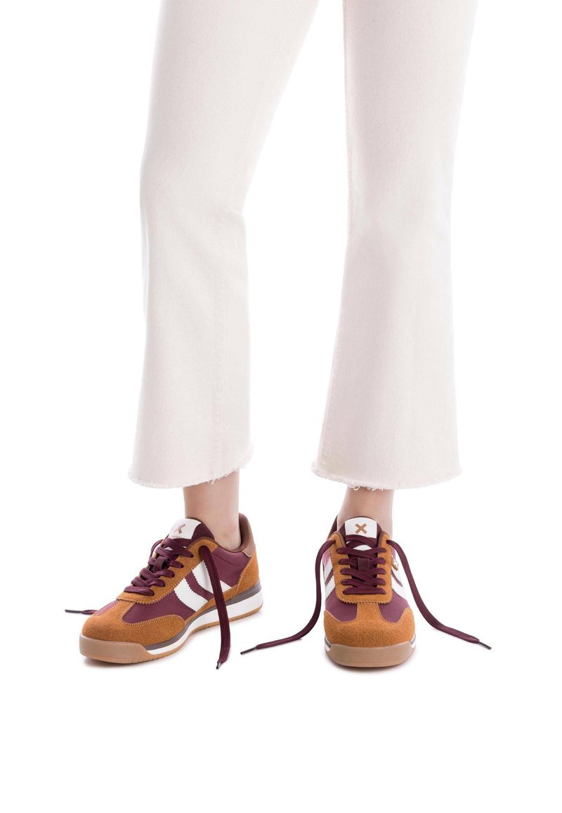Zapatillas bajas de ante marrón y burdeos, con rayas blancas, cordones redondos y suela de goma. Complementan unos jeans blancos de campana.