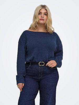 Vrouw met blond haar die een donkerblauwe off-shoulder trui en donkerblauwe jeans met een zwarte riem draagt, poseert tegen een effen witte achtergrond.