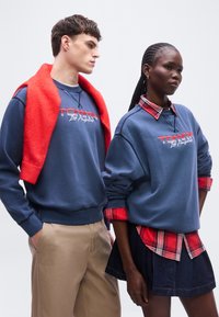 Άνδρας με κόκκινο πουλόβερ περασμένο στους ώμους και γυναίκα με μπλε φούτερ και κόκκινο καρό πουκάμισο σε στρώσεις, ποζάρουν μπροστά σε λευκό φόντο.