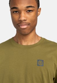 Olivegrünes Baumwoll-T-Shirt mit Rundhalsausschnitt und kurzen Ärmeln, das auf der linken Brustseite mit einem blauen Logo-Patch versehen ist. Glatte Textur.