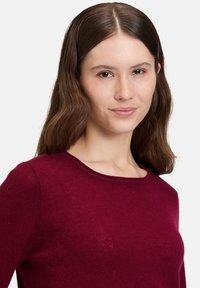 Kvinna med mellanhårig brunt hår som bär en vinröd tröja, vänd framåt med ett neutralt uttryck mot en enfärgad vit bakgrund.