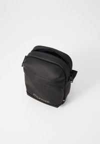 Borsa a tracolla in sintetico nera con parte superiore arrotondata, chiusura con zip e logo "HILFIGER" in rilievo verde oliva. Presenta una tracolla regolabile e un'etichetta del marchio.