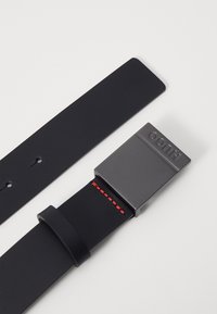 Curea din piele neagră cu o cataramă metalică neagră mată. Prezintă un accent cu cusături roșii pe banda de piele. Design curat, minimalist.