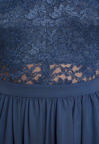 Robe bleu foncé avec un corsage en dentelle orné de motifs floraux, complétée par une jupe plissée et une ceinture lisse. Contraste élégant de textures.