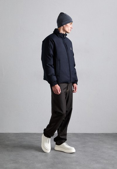 Navy padded jacket with a high collar, black trousers, and white sneakers. Accented with elastic cuffs and a small logo on the chest.