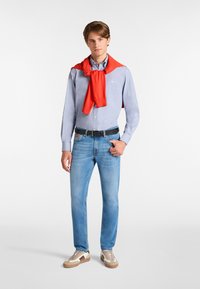 Camicia azzurro chiaro, maglione rosso appoggiato sulle spalle, jeans blu, cintura nera e sneaker beige con dettagli marroni.