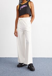 Žena v černém crop topu Puma, bílých kalhotách s volnými nohavicemi a stahovací šňůrkou, a černých platformových teniskách stojí na modrém koberci.