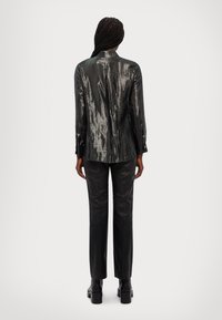 Blazer metallico nero con texture lucida, maniche lunghe e colletto a punta, abbinato a pantaloni in pelle nera aderenti e stivali con tacco spesso.