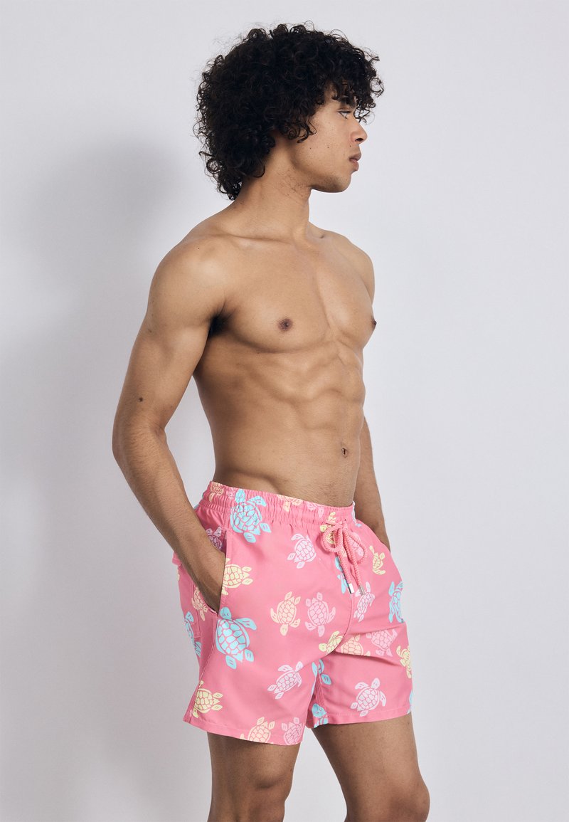 Pantaloni de baie roz cu un model de broaște testoase în galben și albastru, cu un talie elastică și buzunare laterale. Material ușor.