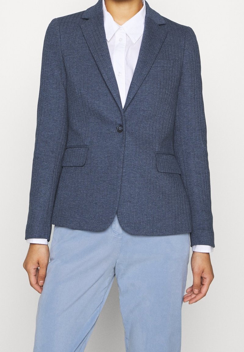 Blazer ajusté bleu marine avec texture chevron, fermeture à un bouton et deux poches avant, associé à une chemise blanche en dessous.
