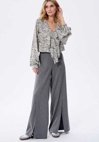 Bloemmotief, lichte blouse met lange mouwen, gecombineerd met hooggeknipte, wijde grijze broek met zijsplitten. Casuele schoenen zichtbaar.