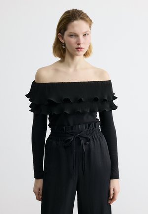 Ung kvinde iført en sort off-shoulder top med flæser og lange ærmer samt højtaljede sorte bukser med snøre.