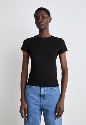 Personne mince portant un t-shirt noir uni à manches courtes et un jean taille haute bleu clair, debout sur un fond blanc.
