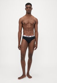 Schwarze Baumwollslips mit einem breiten elastischen Bund, der das "MOSCHINO"-Logo in Rot und Weiß zeigt. Glatte Textur, klassischer Schnitt, minimalistisches Design.