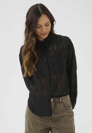 Black sheer button-up shirt with a diamond pattern and long sleeves, worn over olive-green pants with a side pocket.