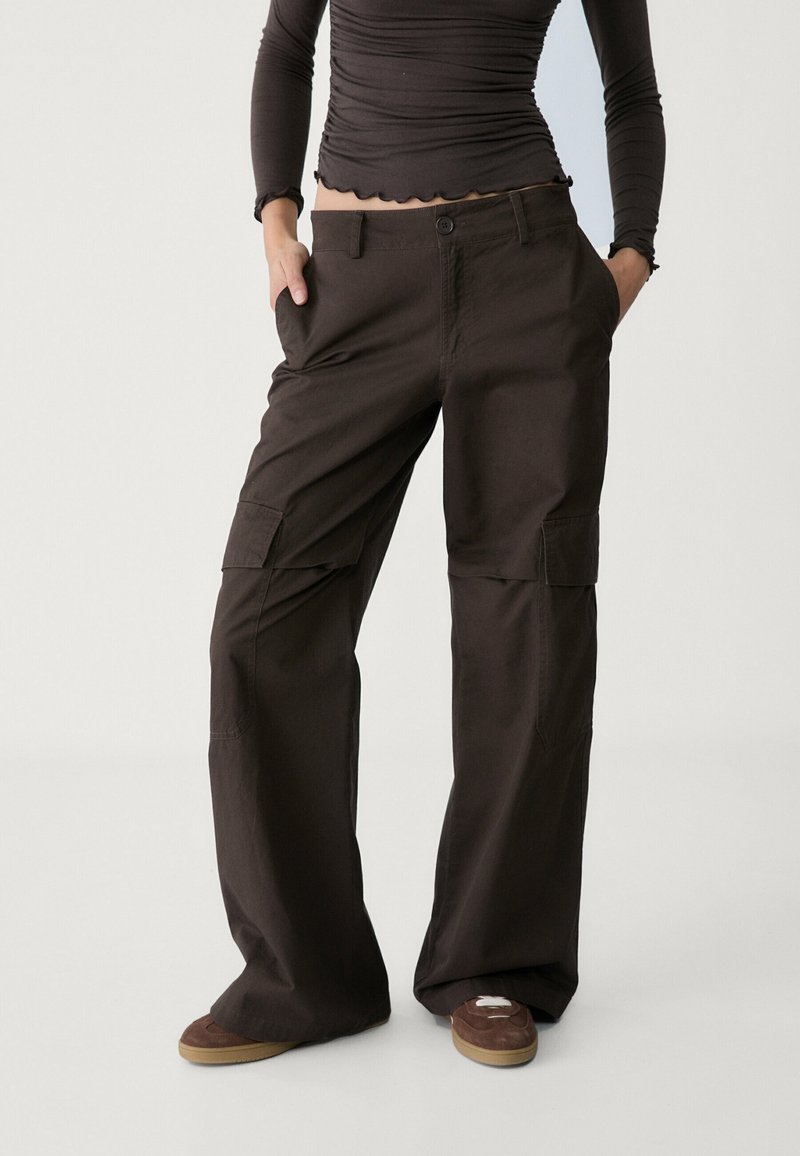 Personne portant un pantalon cargo marron foncé avec les mains dans les poches, associé à un haut ajusté à manches longues et des baskets marron.