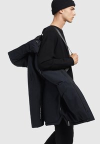 Jachetă izolată bleumarin cu finisaj mat, dotată cu guler captusit, mai multe buzunare cu fermoar și o curea pentru transport ușor.