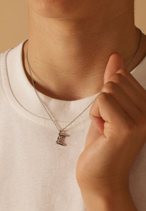 Silver chain necklace with a letter pendant featuring spikes and two decorative gemstones, worn against a light-colored shirt.