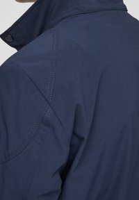 Veste bleu marine avec un col montant, une texture lisse, des coutures visibles et des boutons-pression, présentant un design moderne et une forme ajustée.