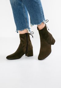 Botas de ante marrón hasta el tobillo con un tacón bajo, que presentan una cremallera lateral, punta afilada y lazos decorativos en la parte posterior.