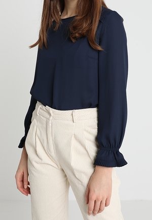 Femme portant un chemisier à manches longues bleu marine foncé avec des poignets volantés et un pantalon en velours côtelé crème taille haute, debout devant un fond uni.