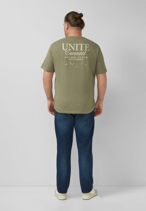 Mężczyzna o brązowych włosach związanych w koczek, ubrany w oliwkowozielony t-shirt z napisem "UNITE Connect", niebieskie dżinsy i białe trampki, stoi zwrócony twarzą do szarej ściany.