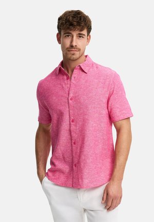Mand iført en kortærmet lyserød skjorte med knapper og krave, stående med den ene hånd i lommen på sine hvide bukser.