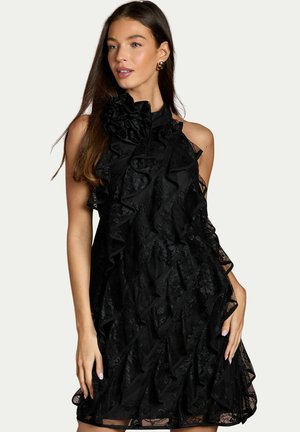 Frau mit langen braunen Haaren, die ein ärmelloses schwarzes Spitzenkleid mit Rüschen und goldene Creolen trägt, posiert mit einer Hand in der Hüfte.