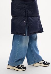 Giacca puffer blu marina con finitura lucida, dotata di bottoni a pressione argentati, abbinata a jeans a gamba larga blu chiaro e scarpe da ginnastica nere con dettagli bianchi.