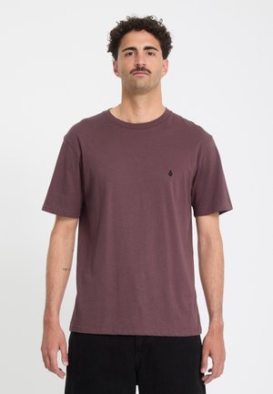 Vuntsidega mees, kes kannab ühevärvilist pruuni T-särki ja musti pükse, seisab ühevärvilise valge tausta vastu.