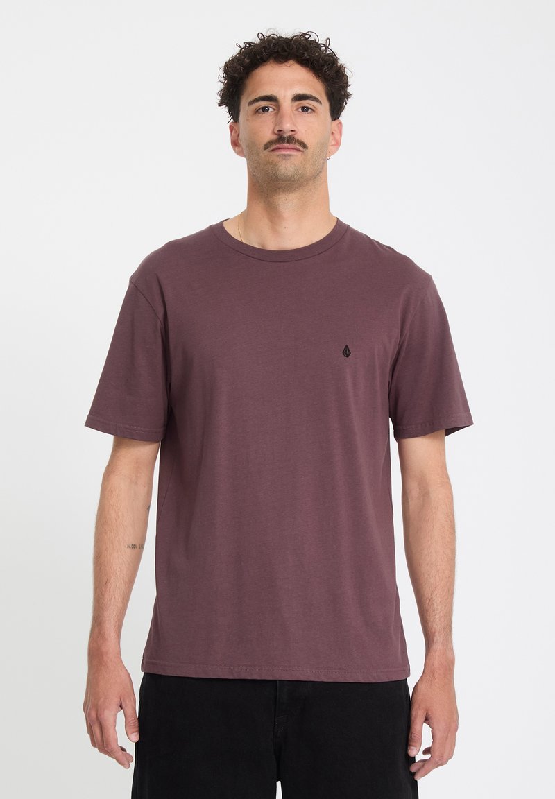 Άντρας με μουστάκι που φοράει απλό καφέ T-shirt και μαύρο παντελόνι, στέκεται μπροστά σε απλό λευκό φόντο.