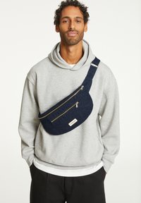 Sac bandoulière en tissu bleu avec deux compartiments zippés, forme incurvée et bandoulière ajustable, porté sur un sweat à capuche gris.